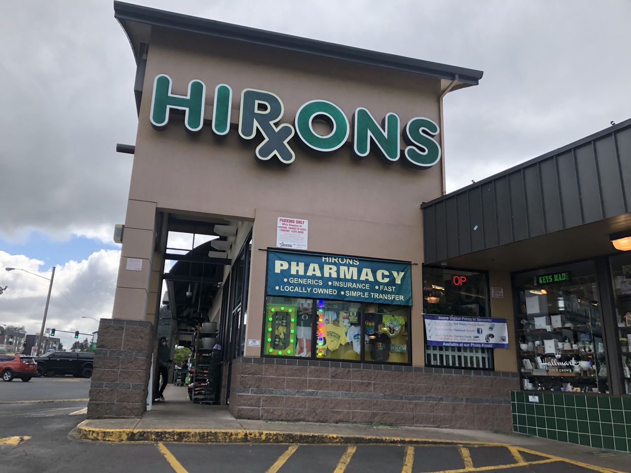 A tan building with green lettering on the front spelling out "Hirons" with a pharmacy "Rx" symbol used for the "R". Below the logo is a window with the words "pharmacy" and filled with lights and green and yellow Oregon Ducks gear.