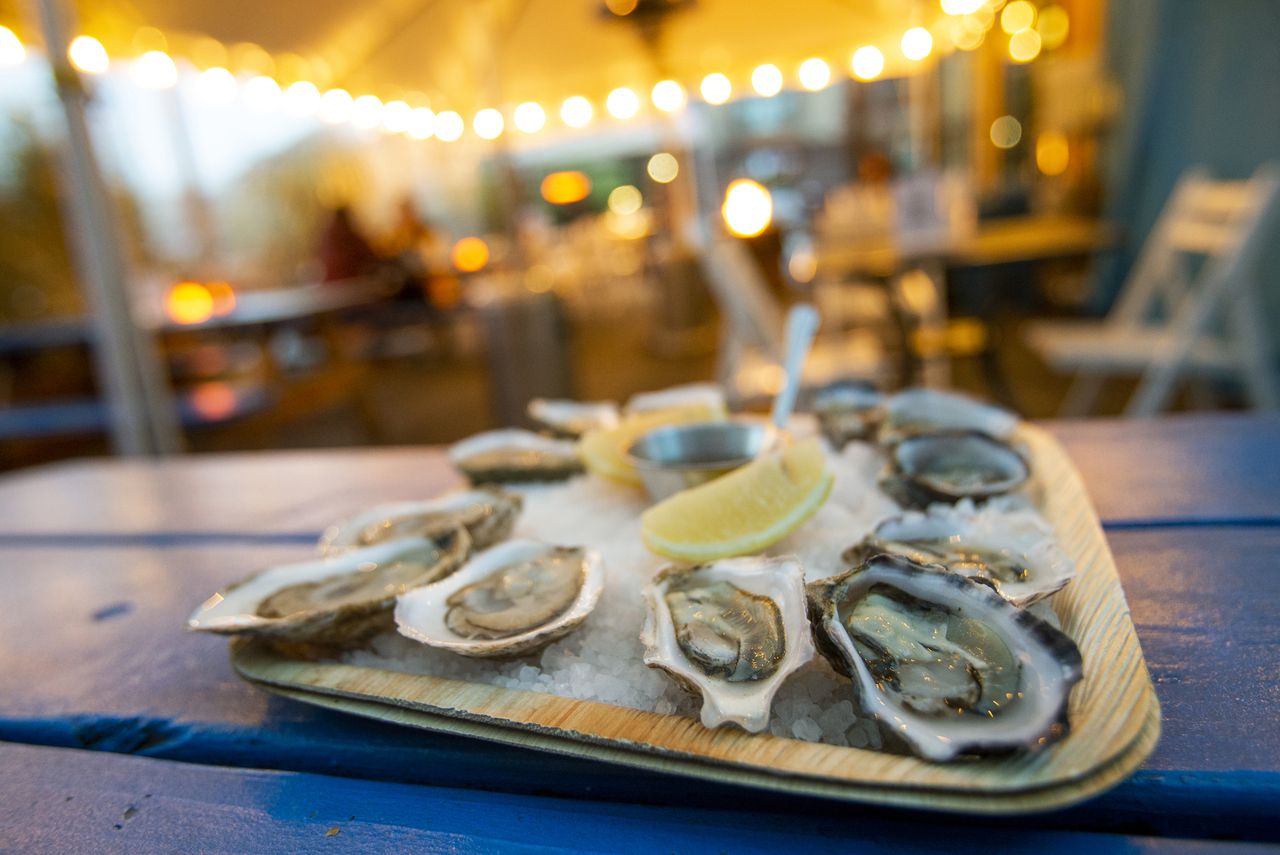 A plate of a dozen oysters, sitting on a blue benchtop, with an outdoor dining area in the background. The plate is covered in rock salt with the oysters arranged around the center, where a cup of sauce and two lemon wedges are located